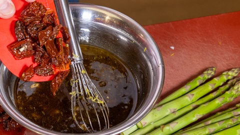Kleingeschnittene getrocknete Tomaten werden mit einer Hand von einem roten Teigschaber in eine Schüssel mit bräunlicher Flüssigkeit gegeben.