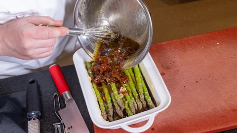 Grüner Spargel in einer weißen Auflaufform wird mit einer dunklen Flüssigkeit mit Tomaten drin übergossen
