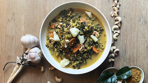 Grünkohlcurry in einem weißen Suppenteller auf Holztisch, daneben Zitrone, Nüsse, Käse und dunkle Blätter