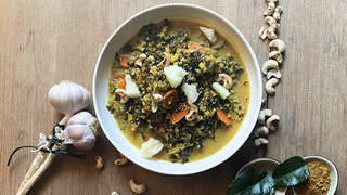 Grünkohlcurry in einem weißen Suppenteller auf Holztisch, daneben Zitrone, Nüsse, Käse und dunkle Blätter