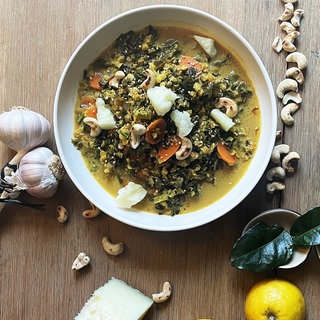 Grünkohlcurry in einem weißen Suppenteller auf Holztisch, daneben Zitrone, Nüsse, Käse und dunkle Blätter