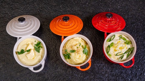 Drei kleine Töpfe mit schräg angelehnten Deckeln stehen auf einer dunklen Unterlage. Die Farben der Töpfe sind grau, orange und rot. In den Töpfen befindet sich je ein Kartoffelpüree mit grünen Kräutern und Lauchzwiebeln dekoriert.