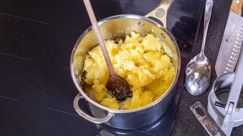 Ein Topf mit einem Stil steht auf einem Herd, im Topf sind halb zerstampfte Kartoffeln und ein Kochlöffel zum Umrühren des Kartoffelpürees