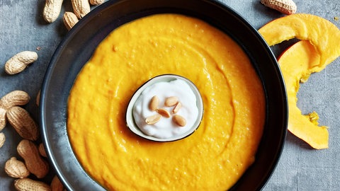Rezept Kürbis-Erdnuss-Suppe mit Joghurt Dip: Kürbissuppe in einem dunklen Teller mit einer kleinen, hellen Schale mit Joghurt-Dip in der Mitte. Drumherum liegen Erdnüsse in Schale. 