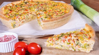 Rezept Lauchkuchen: Der Lauchkuchen ist angerichtet auf einem Holzbrett. Daneben liegt ein Lauch, kleine Tomaten und ein Schälchen mit Schinkenwürfeln.