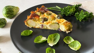 schwarzer Teller mit einem Stück Rosenkohl-Quiche, drumherum Rosenkohlblätter