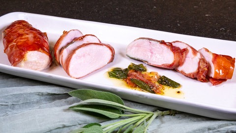 Rezept für Saltimbocca vom Hähnchen: Mehrere Scheiben von Saltimbocca aus rosafarbenem, saftigem Hähnchenfleisch liegen aufgeschnitten auf einem länglichen Teller, daneben sind Stängel von Salbei zu sehen.