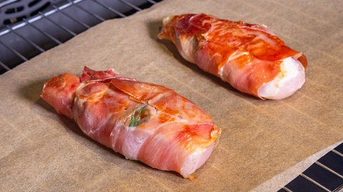 Rezept für Saltimbocca vom Hähnchen: Die mit Schinken umwickelten Hähnchen-Stücke liegen in einem Ofen auf Backpapier und werden gegart.