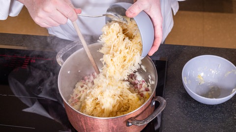 Ein Koch kippt Sauerkraut in einen Topf und rührt mit deinem Kochlöffel um. Daneben steht eine hellgraue leere Schüssel. 