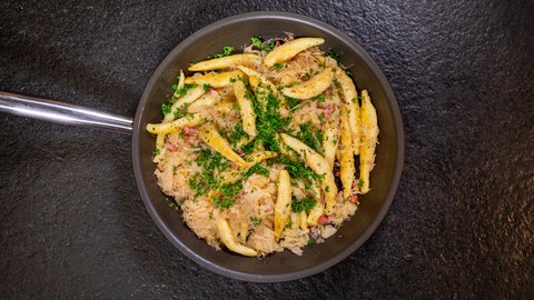 In einer Pfanne sind Schupfnudeln mit Sauerkraut und Speck auf einer schwarzen Arbeitsplatte angerichtet.