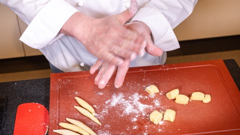 Ein Koch formt mit den Händen aus einem Kartoffelteig Schupfnudeln und legt sie auf ein rotes Küchenbrett für Schupfnudeln mit Sauerkraut und Speck.