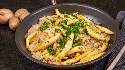 Auf einer schwarzen Arbeitsplatte ist eine Pfanne mit Schupfnudeln, Sauerkraut und Speck angerichtet. Daneben lieben 4 kleine Kartoffeln und ein Kochlöffel.