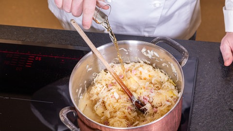 Ein Koch kippt Apfelsaft in einen kupferfarbenen Topf mit Sauerkraut und Speck.