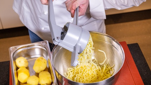 Ein koch reibt in eine Schüssel gekochte Kartoffeln. Daneben liegt eine Metallschüssel mit noch ganzen Kartoffeln für Schupfnudeln mit Sauerkraut und Speck.