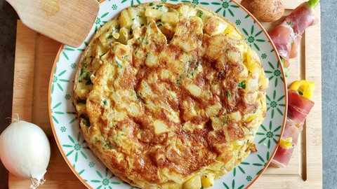 Tortilla auf einem hellen Teller, daneben eine Zwiebel, ein Holzschaber und Gemüse in Schinken gewickelt