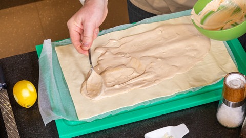 Für das Rezept einer Tomaten-Tarte wird eine Creme aus Schmand auf einem ausgerollten Blätterteig verteilt.