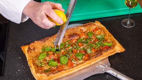 Eine fertig gebackene Tomaten-Tarte wird mit Basilikum belegt und mit geriebener Zitrone beträufelt.
