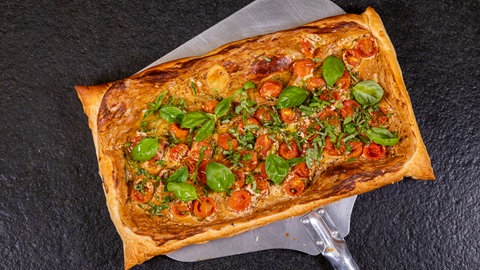 Eine Tomaten-Tarte aus Blätterteig mit Kirschtomaten und Basilikum liegt fertig gebacken auf einem Backschieber.