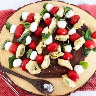 Rezept für Tortellini-Spieße: Spieße mit Tortellini, Tomaten, Basilikum auf einer Holzplatte. Daneben ein Holzlöffel mit grobem Salz