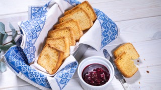 In einer rechteckigen Box, die mit einem blau gemusterten Tuch ausgelegt ist, liegen sechs goldgelbe Zwiebackscheiben. Daneben steht ein weißes Schälchen mit einer roten Marmelade und danebem liegt ein Buttermesser.