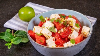 Salat aus Wassermelone, Feta, Minze, Pinienkernen und Limetten-Dressing ist angerichtet in einer blauen Schüssel.