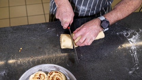 Der aufgerollte Hefeteig wird in Scheiben geschnitten. Im Vordergrund liegen Zimtschnecken in einer runden Backform.