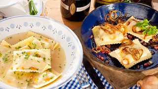 Maultaschen stehen auf einem gedeckten Tisch: auf der linken Seite in einem Teller mit Brühe, auf der rechten Seite in einer kleinen Pfanne, geschmälzt mit gebratenen Zwiebelringen. Dahinter zwei Weinflaschen mit Trollinger und Riesling. In SWR1 verrät Anton Schmaus, der Koch der deutschen Fußball-Nationalmannschaft, sein ganz persönliches Maultaschenrezept - zum ersten Mal! Und Nationalspieler Jamal Musiala kommt auch aus der Maultaschen-Deckung, mit einem überraschenden Tipp!