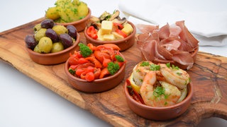 Auf einem Brett stehen sechs Schälchen mit verschiedenen spanischen Tapas: Oliven, Garnelen, Kartoffeln, Schinken, Paprika. Ob Garnelen, Oliven, Kartoffeln, getrocknete Tomaten ... die Möglichkeiten für Tapas sind grenzenlos. SWR1 Pfännle-Koch Eberhard Braun liefert euch Tapas-Inspirationen.