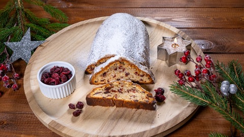 Ein nach SWR4 Rezept fertig gebackener Stollen mit Cranberry und Marzipan angeschnitten auf einem runden Holzbrett mit einer brennenden Kerze in Sternform und einer Schale getrockneter Cranberrys