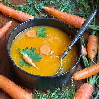 Ein schwarzer Topf mit Karottensuppe steht auf einem Holztisch. Um den Topf herum liegen Karotten.