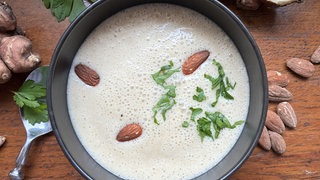 Mandelsuppe in einer schwarzen Schale auf einem Holztisch. Um die Schale herum liegen Mandeln, Topinambur und ein Löffel.