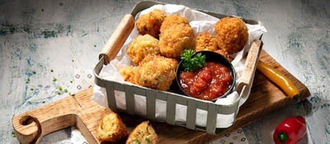 Auf einem Tisch steht ein Glasbehälter mit einer Portion Käsespätzle-Arancini. Daneben ein Schälchen mit einer Salsa-Sauce.