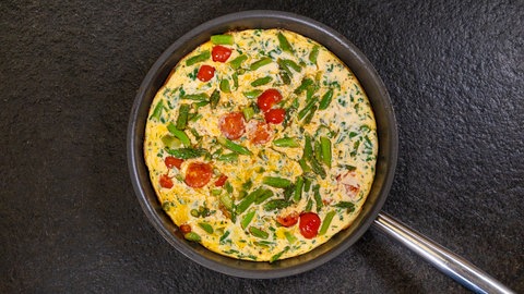 Eine Pfanne mit einem Omelett aus Eiern, grünem Spargel, Kirschtomaten, Sahne und Parmesan auf einer schwarzen Arbeitsplatte.