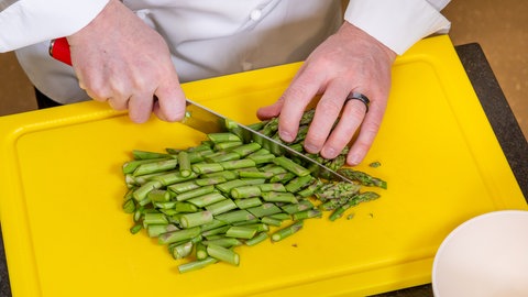 Ein Koch schneidet auf einem gelben Schneidebrett Stangen vom grünen Spargel in kleine Stücke.