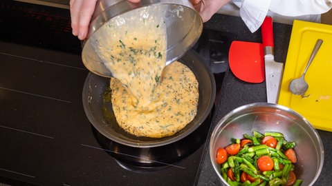 Ein Koch schütten eine Omelett-Masse aus Eiern, Sahne, Parmesan und Petersilie in eine heiße Pfanne und daneben steht eine Schüssel mit angebratenem grünen Spargel und Kirschtomaten.