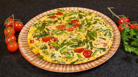 Ein Omelett aus grünem Spargel auf einem Teller serviert und mit frischen Kirschtomaten auf einer schwarzen Arbeitsplatte angerichtet.