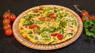 Ein Omelett aus grünem Spargel auf einem Teller serviert und mit frischen Kirschtomaten auf einer schwarzen Arbeitsplatte angerichtet.