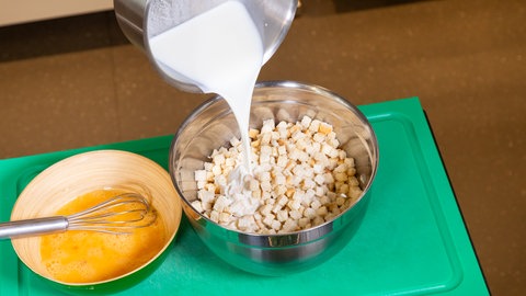 Ein Koch kippt einen Topf Milch in eine Schüssel mit Brötchenwürfeln. Daneben streht eine Schüssel mit verquirlten Eiern.