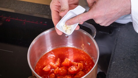Ein Koch gibt gehackten Knoblauch aus einer kleinen Schüssel in einen Topf mit Tomatensauce und halbierten Cherrytomaten.