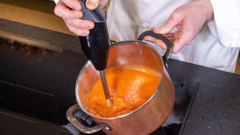 Ein Koch püriert mit dem Pürierstab eine Tomatensauce in einem Topf und hält ihn schräg von sich weg.