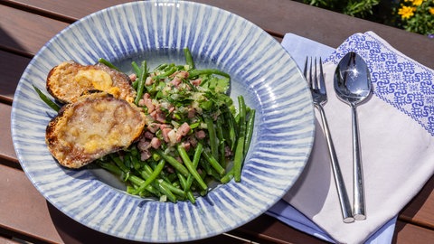 Grüner Bohnensalat und geröstetes Brot mit Bergkäse überbacken auf einem blaugemusterten Teller angerichtet und mit einer Serviette und Besteck daneben dekoriert.