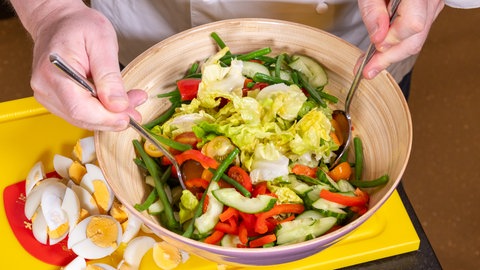 Ein Koch bereitet einen Nizza-Salat zu und rührt in einer Schüssel Kopfsalat, grüne Bohnen, Paprika, Gurken in dem Dressing zusammen. Daneben liegen aufgeschnittene hartgekochte Eier.