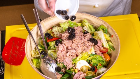 Ein Koch bereitet einen Nizza-Salat zu und gibt Oliven in die Schüssel mit dem Salat aus Ei und Thunfisch, Paprika, Kirschtomaten und einem leckeren Dressing. 