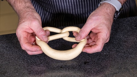 Zwei Hände halten eine noch frisch geformte und rohe Brezel über einer schwarzen Arbeitsfläche in die Kamera.