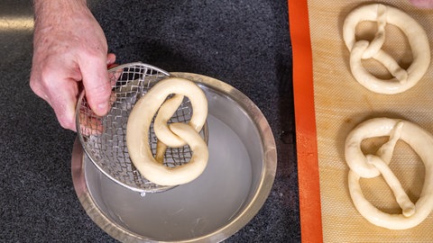 Eine Hand hält einen zu einer Brezel geformten Teig mit einem Sieb über eine Metallschüssel mit Natronwasser, rechts davon liegen zwei ungebackene Brezeln auf einem Blech