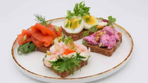 Vier verschiedene Smørrebrød auf einem Teller. Smörrebröd ist mehr als ein belegtes Brot: Es sind kleine Kunstwerke, die liebevoll angerichtet werden. Die reichlich belegten Butterbrote sind in Dänemark ein traditionelles Mittagessen. Grundlage ist gebuttertes Roggenbrot.