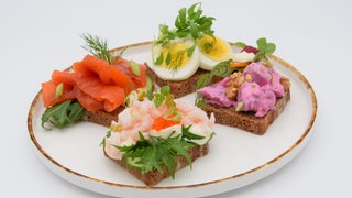Vier verschiedene Smørrebrød auf einem Teller. Smörrebröd ist mehr als ein belegtes Brot: Es sind kleine Kunstwerke, die liebevoll angerichtet werden. Die reichlich belegten Butterbrote sind in Dänemark ein traditionelles Mittagessen. Grundlage ist gebuttertes Roggenbrot.