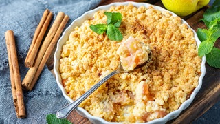 Rezept Apple Crumble mit Zimt: In einer weißen Schale ist Apple Crumble (gebackene Äpfel mit Streusel) angerichtet. Daneben liegen mehrere Zimtstangen und ein paar Äpfel.