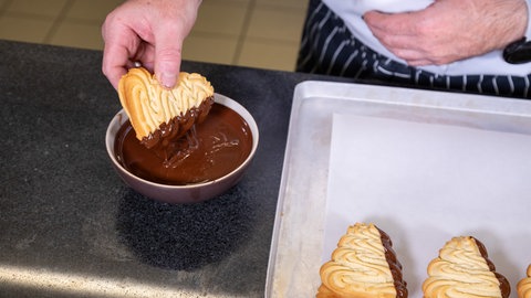 Eine Hand tunkt die fertig gebackenen wellenförmigen Teigstücke seitlich in flüssige dunkle Schokolade, die in einer Schüssel neben einem Bleck ist