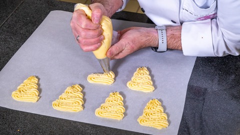 Zwei Hände füllen Teig in Wellenform unten breit oben scmal aus einer Spritztülle auf eine Silikonmatte